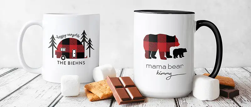 A pair of white mugs featureing custom camping designs are shown with s.mores fixing
